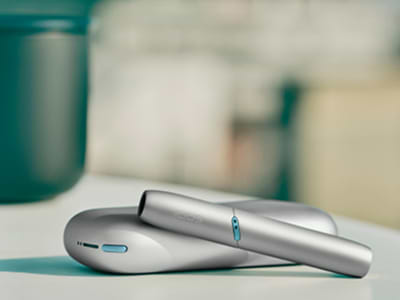 Man holding IQOS Originals Duo device in Turquoise in one hand and a mobile phone in the other hand.