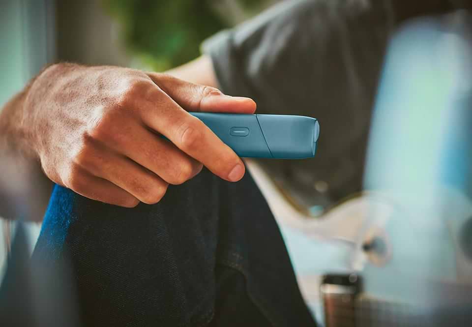 Turquoise IQOS Originals One device on a desk next to a tea cup.	 	 	
