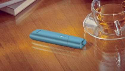 Turquoise IQOS Originals One device on a desk next to a tea cup.