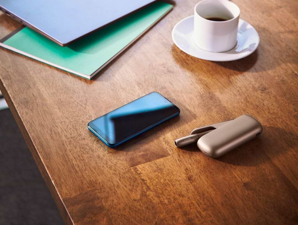 A gold IQOS, a mobile and a cup of coffee on a wooden table.