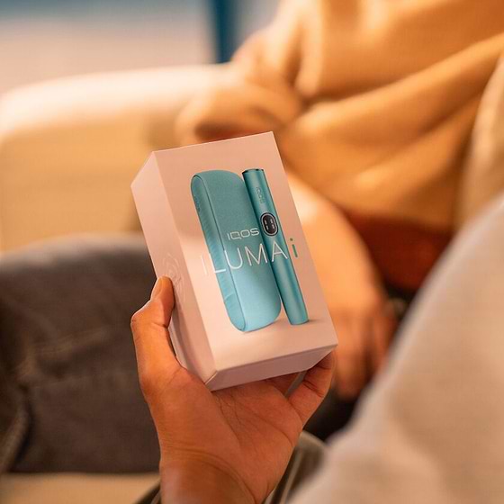 A woman holding an IQOS ILUMA device while looking at a laptop.