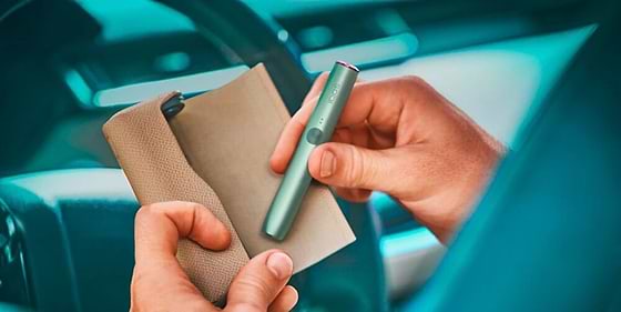 An IQOS device held by two hands in front of a car steering wheel.