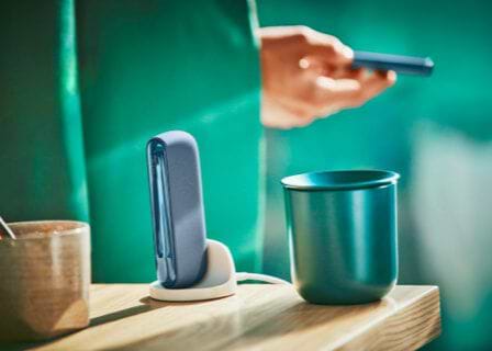 A man holds an IQOS ILUMA mid azure blue device, with a holder and cup on the table.