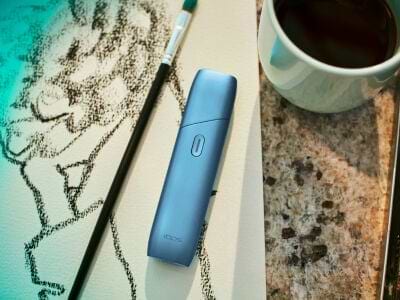 Turquoise IQOS Originals One device on a painting, next to a coffee cup.	 	 	