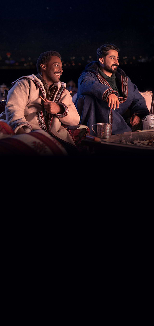 رجلان يجلسان في الهواء الطلق ليلاً، يرتديان عباءات تقليدية دافئة، يبتسمان ويستمتعان بجلسة مريحة مع وسائد مزخرفة وطاولة تحتوي على شاي ووجبات خفيفة.