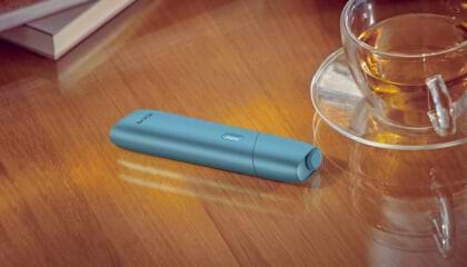 Turquoise IQOS Originals One device on a desk next to a tea cup.	 	 	