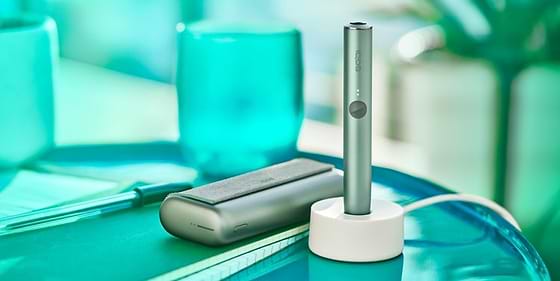 An IQOS ILUMA device in a charging station with a Pocket Charger on a table.