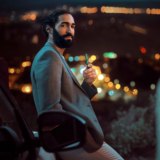 Confident man in a gray suit holding an IQOS device at night with blurred city lights in the background.