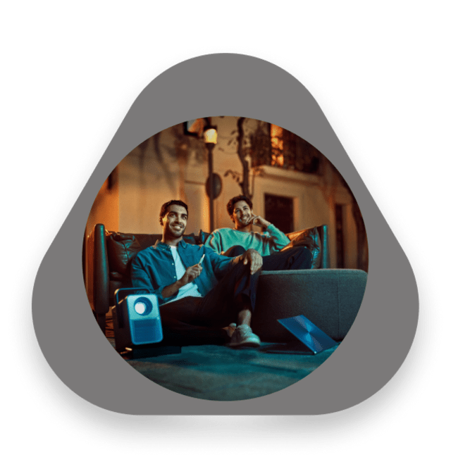 رجلان شابان يجلسان على الأرائك في الهواء الطلق ليلاً، يبتسمان ويسترخيان مع جهاز آيكوس وجهاز عرض محمول بجانبهما.