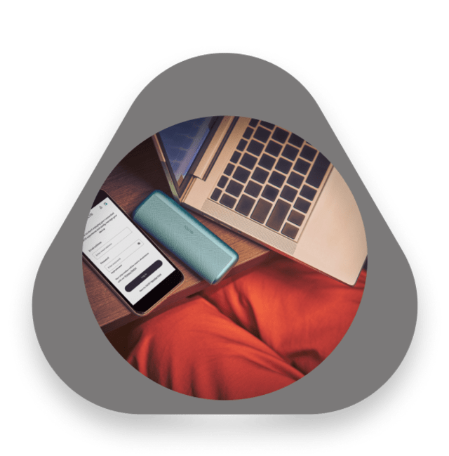 هاتف ذكي مع تطبيق آيكوس مفتوح بجانب جهاز آيكوس باللون الفيروزي وجهاز كمبيوتر محمول على طاولة خشبية، وشخص يرتدي بنطالًا أحمر يجلس بالقرب.