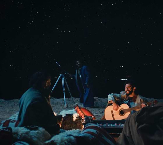 Group of friends sitting outdoors at night under a starry sky, one playing guitar by the fire, another near a telescope, enjoying a cozy gathering.