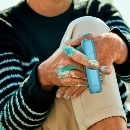 Hands with paint holding a turquoise IQOS Originals One heated tobacco device.	 	 	