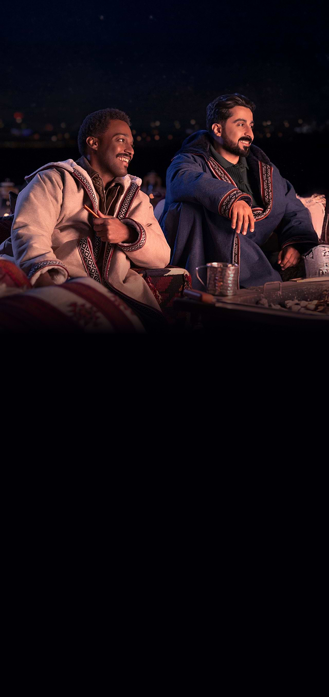Two men sitting outdoors at night, dressed in traditional warm cloaks, smiling and enjoying a cozy gathering with patterned cushions and a table with tea and snacks.