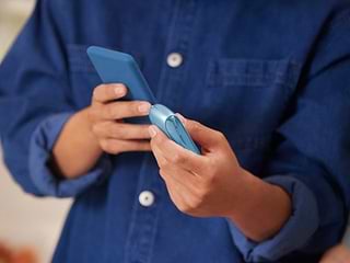 Hands holding turquoise IQOS Originals Duo device and mobile phone. 	 	 	